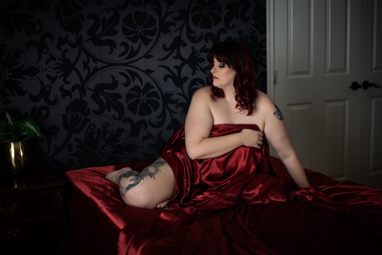 A woman partially draped in red satin sheets sits on a bed, showing her thigh tattoo, during a boudoir session by Leslee Shaw Photography in Sweetwater, Texas.”