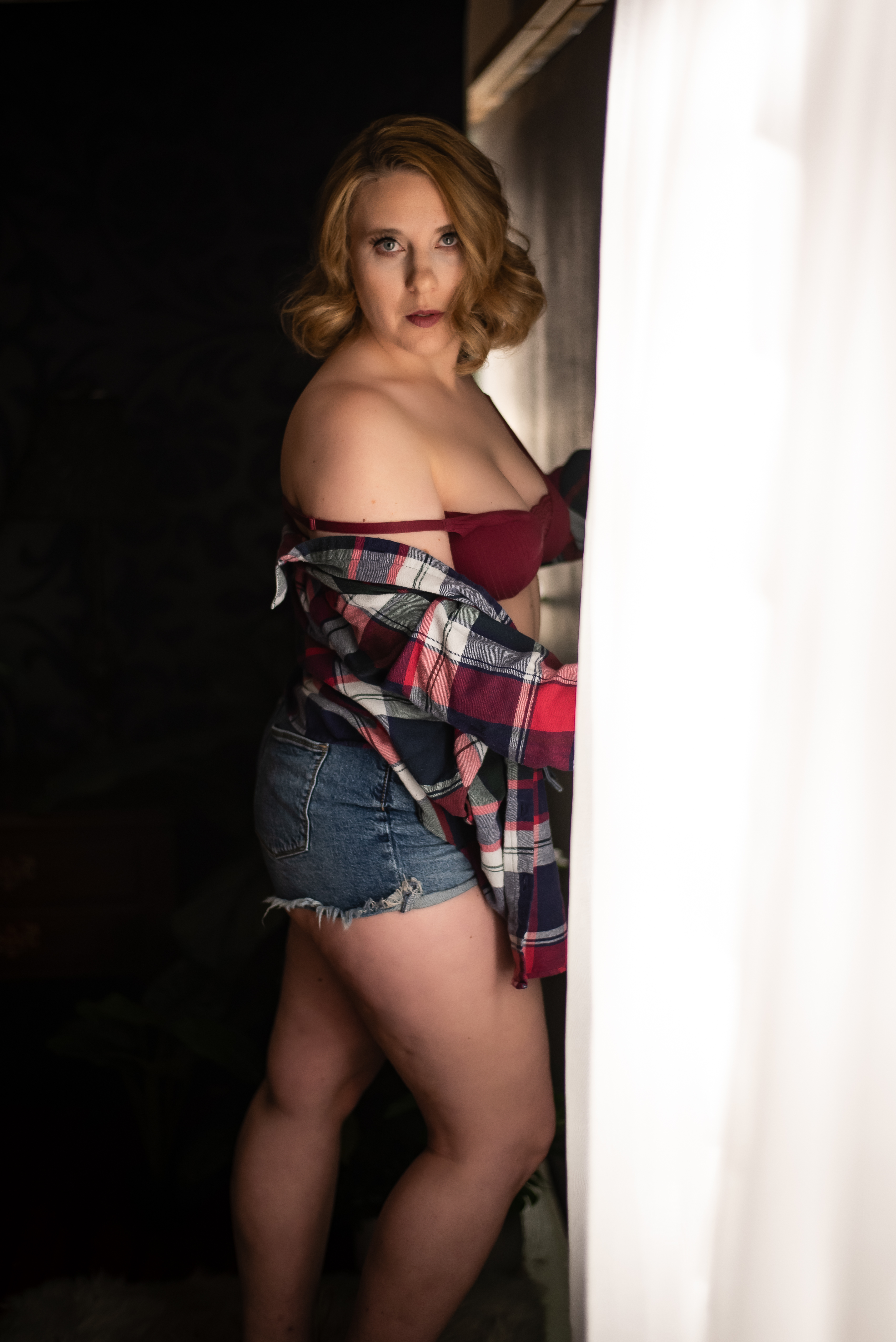 A woman wearing a burgundy bra, flannel shirt, and denim shorts stands by a window, captured by Leslee Shaw Photography in Sweetwater, Texas, showcasing a confident boudoir pose.