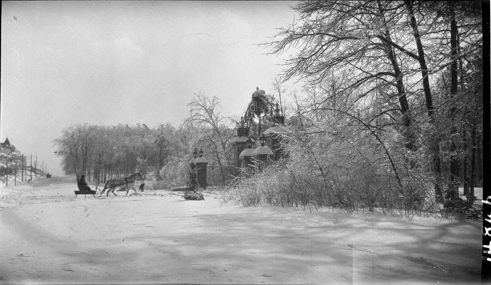 A historic black-and-white winter photograph of High Park in Toronto, showing a horse pulling a sleigh across snow-covered ground near an ornate park entrance. Photo Courtesy: City of Toronto
