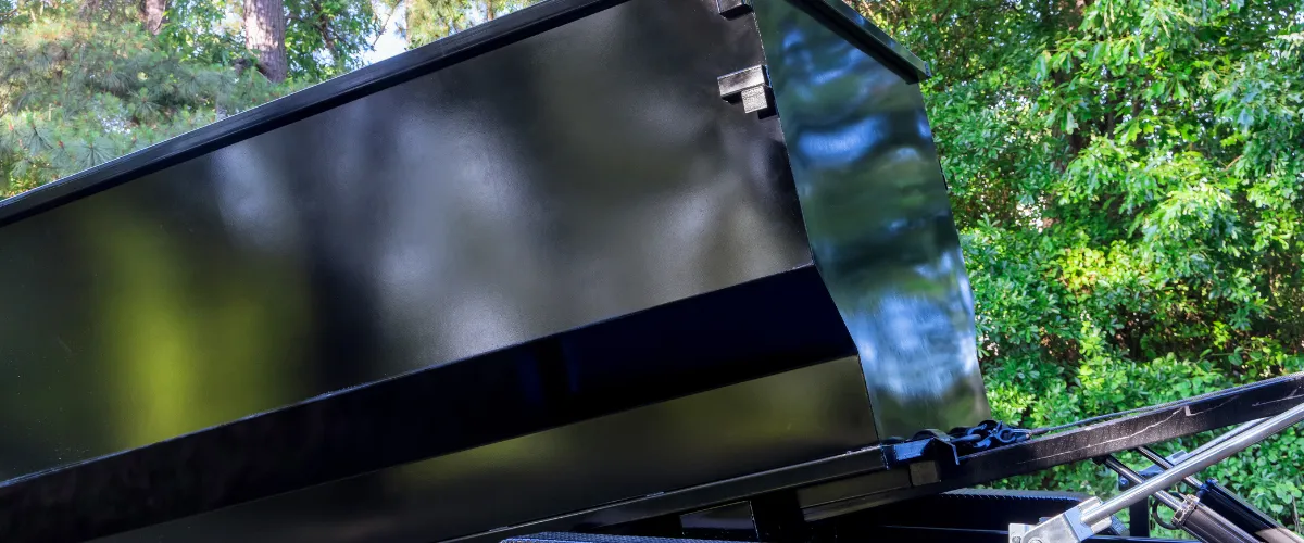 Raised black dumpster trailer surrounded by green trees Raised black dumpster trailer surrounded by green trees