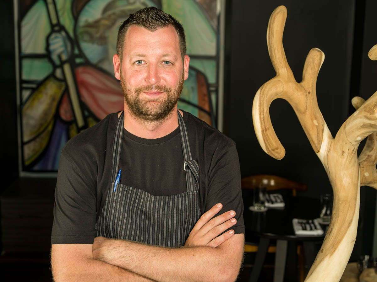 Confident bearded chef in striped apron standing in front of stained glass artwork and wood sculpture decor
