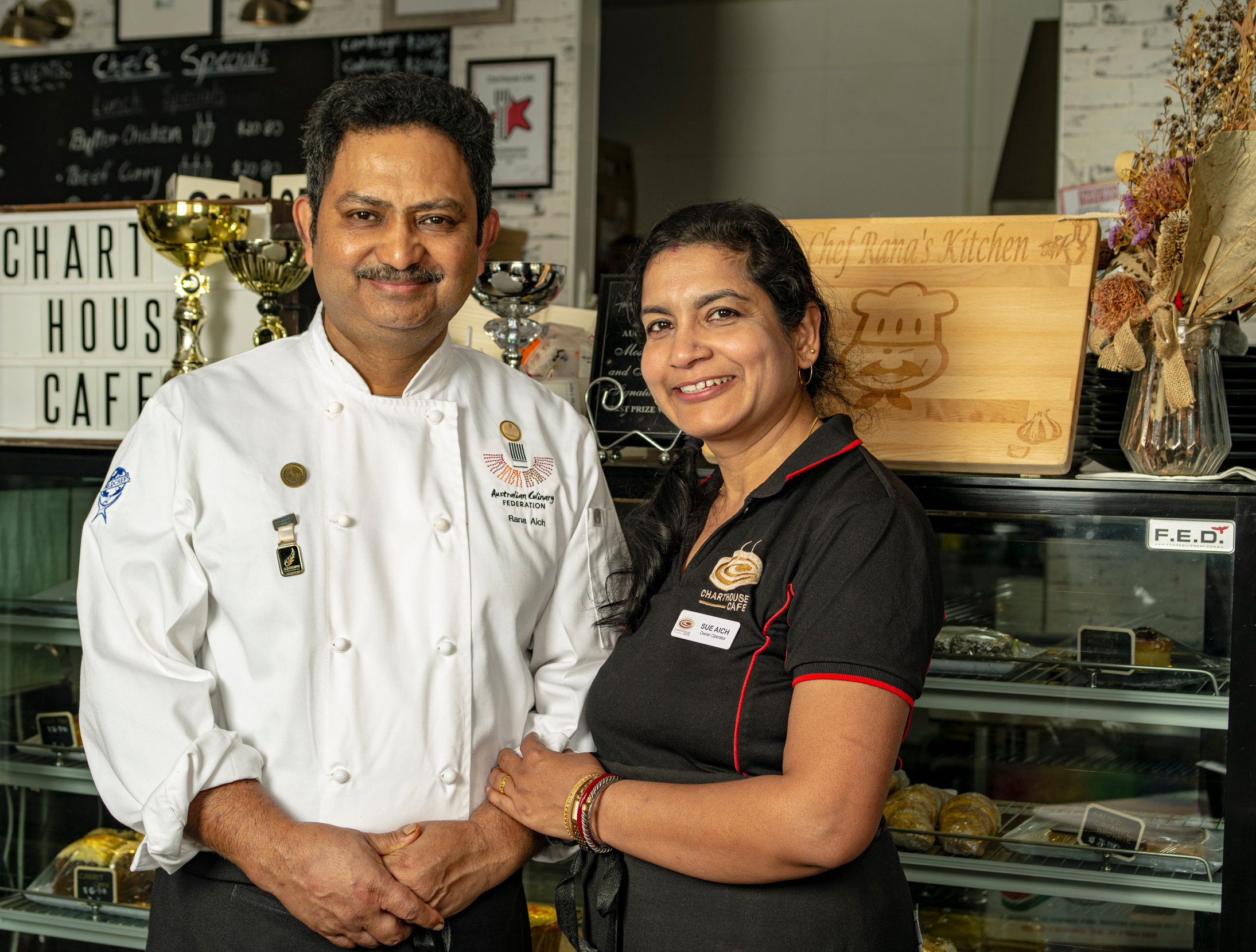 Married chef duo posing in front of their café counter with baked goods and award signs