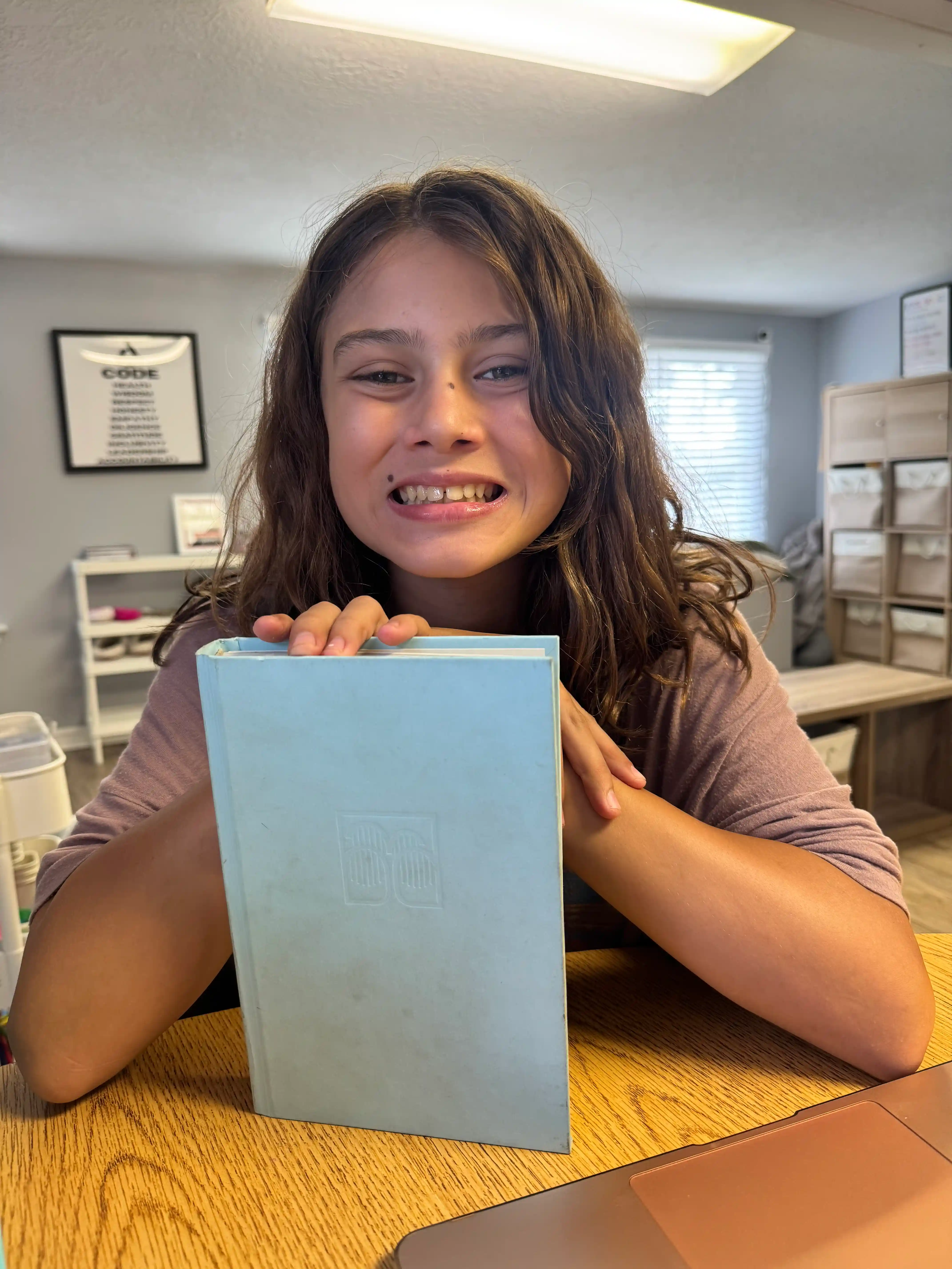 young child holding a book and smiling, sitting at a table in a school setting
