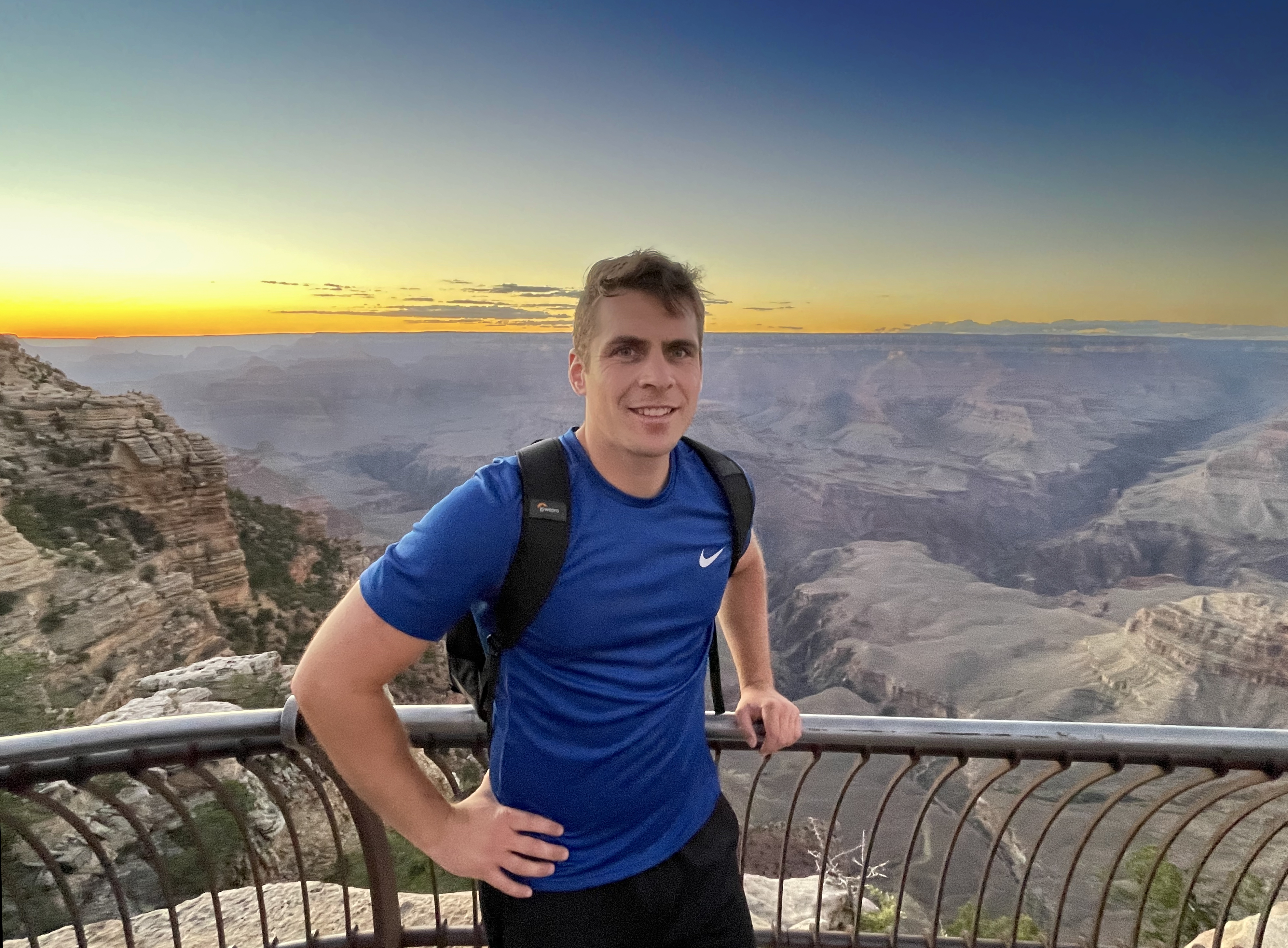 Matthew McNab, man standing in front of sunset at the Grand Canyon National Park smiling at the camera. He has a backpack on his back and his holding the railing with his left arm.