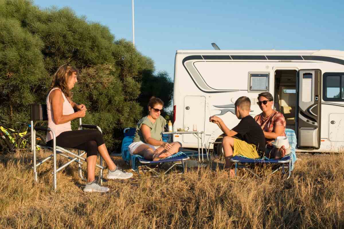 A group of people relaxing outdoors near a white RV van, sitting on camping chairs and a portable bed, with trees and a clear blue sky in the background A group of people relaxing outdoors near a white RV van, sitting on camping chairs and a portable bed, with trees and a clear blue sky in the background