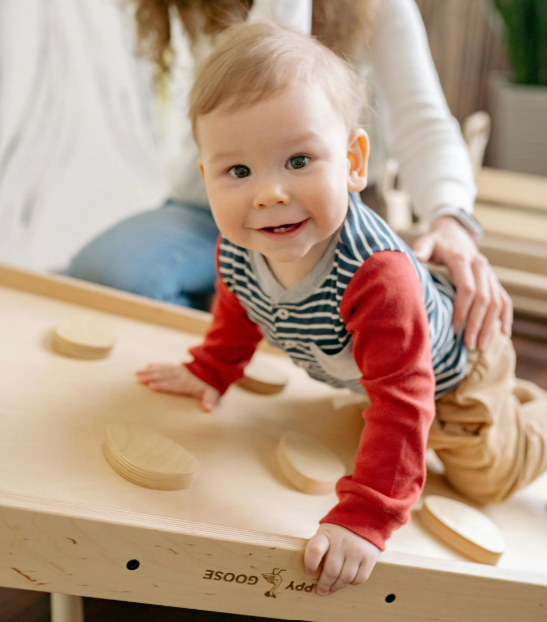 A woman is sitting next to a baby who is crawling on a wooden table.