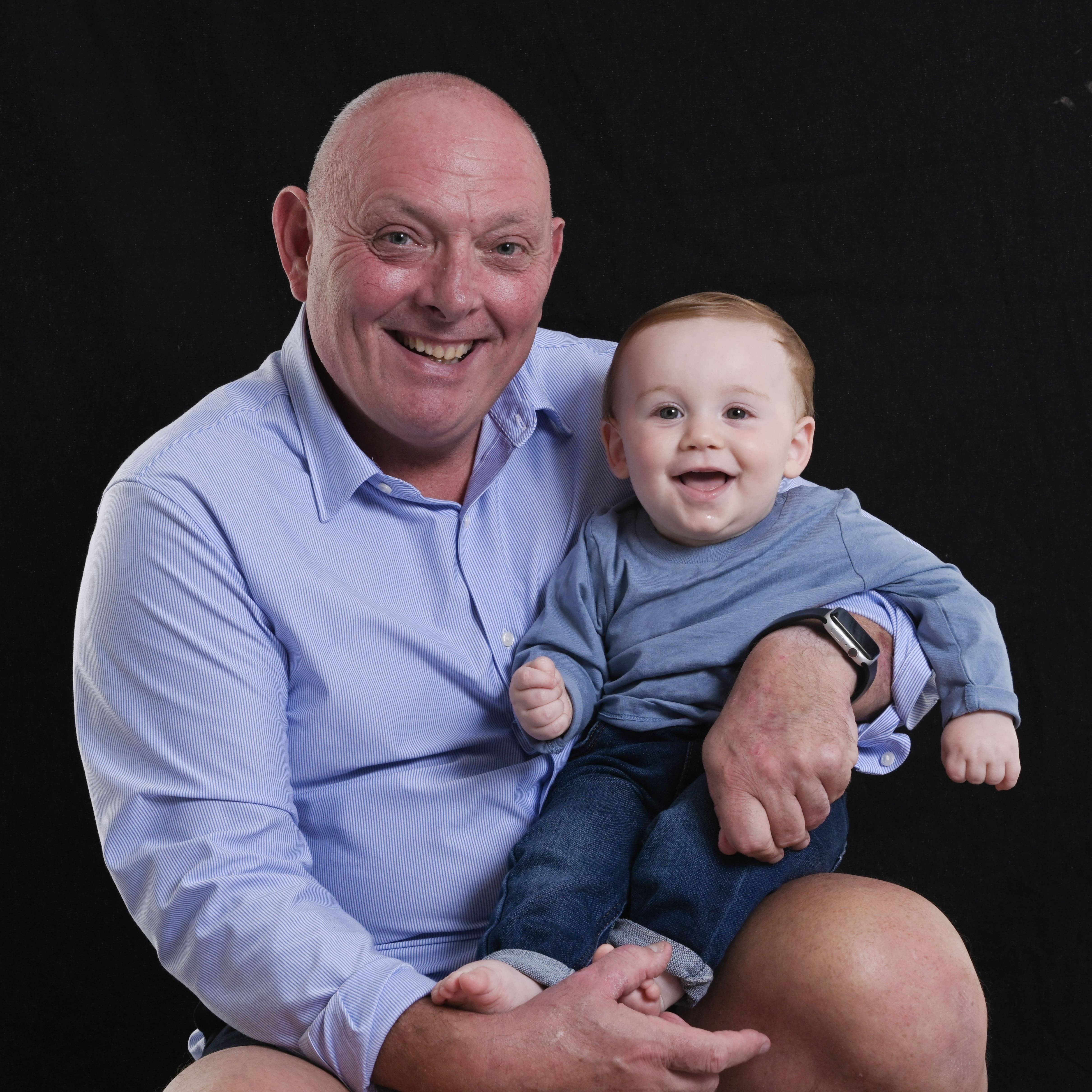 Photo of dad and son smiling at the camera. Dad is holding is young child close to him.