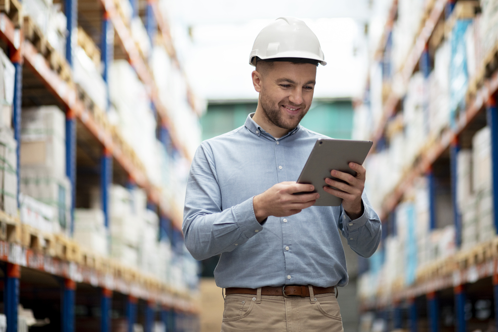 Homem usando chapéu em uma indústria, olhando para seu tablet enquanto analisa seu e-commerce; ao fundo, um grande salão com pallets e produtos empilhados. Homem usando chapéu em uma indústria, olhando para seu tablet enquanto analisa seu e-commerce; ao fundo, um grande salão com pallets e produtos empilhados.