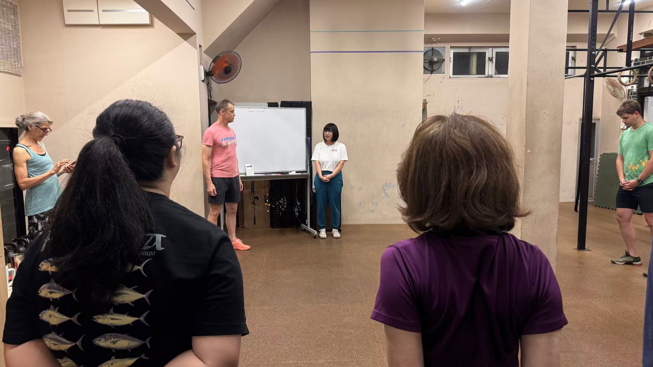Coach leading a CrossFit Kyoto class with participants seen from behind inside the gym. Coach leading a CrossFit Kyoto class with participants seen from behind inside the gym.