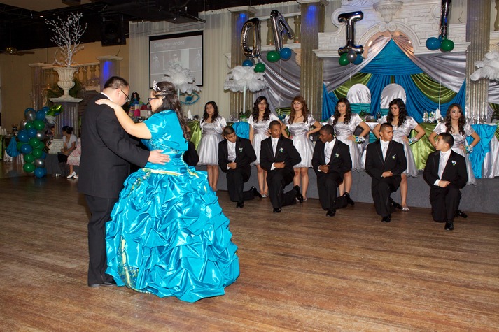 Quinceañera in a bright blue gown dancing with her father on the dance floor, with the court of honor standing and kneeling in the background during the quinceañera program.