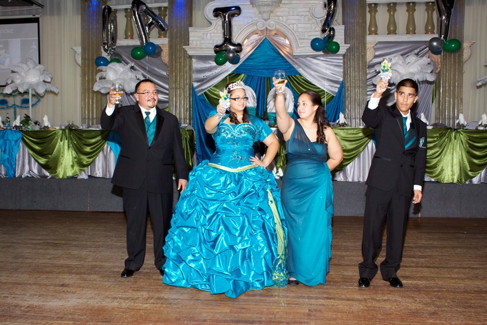 Quinceañera in a bright blue gown raising a toast with her family during the quinceañera program, standing on the dance floor in front of a decorated backdrop.