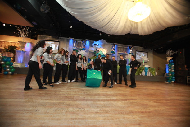 Quinceañera surrounded by her court and family as she opens a surprise gift during the quinceañera program on the dance floor.