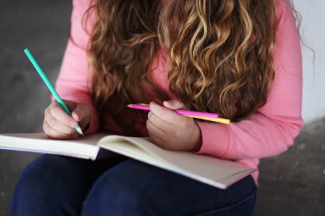 A young girl in a pink sweater holding two colored pens while writing in an open notebook. This image highlights the importance of not relying solely on memory for a quinceañera speech, emphasizing the need to jot down key points to avoid forgetting important things during the speech.