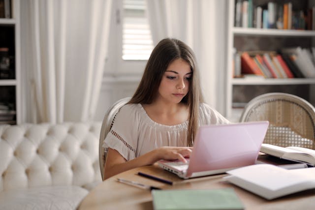 A focused young woman in a white blouse working on a pink laptop at a home study desk surrounded by notebooks and papers. The scene underscores the importance of practicing a quinceañera speech in advance to ensure confidence and fluency during the actual event.