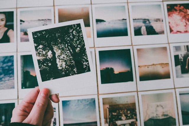 A hand holding a polaroid photo over a table filled with other printed polaroid memories. The image conveys the idea of creating a personalized memory book for your quinceañera court, filled with special moments and memories, as a thoughtful and unique gift to make them feel cherished.