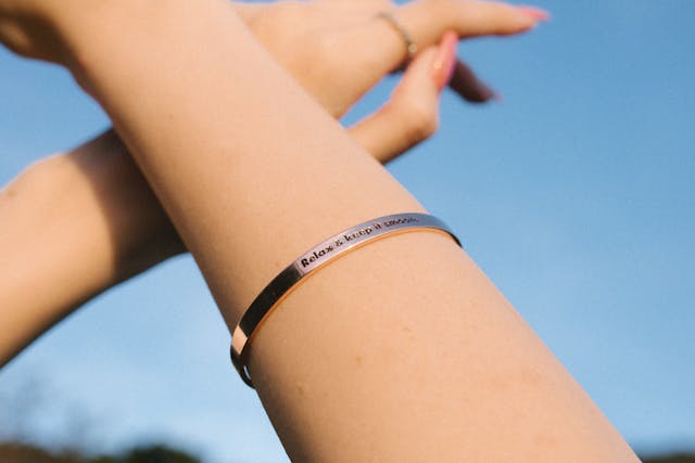 A close-up of a wrist wearing a sleek, personalized bracelet engraved with a message, against a clear blue sky. The image suggests the thoughtful idea of gifting your quinceañera court members personalized jewelry, such as engraved bracelets or necklaces, as a meaningful keepsake to commemorate the special occasion.