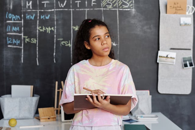 A young girl holding a notebook and pencil, standing in front of a large chalkboard with a weekly schedule drawn on it. The image highlights the importance of planning and scheduling rehearsals with a quinceañera choreographer, ensuring that there is enough time to practice and perfect the dance routines before the big day.