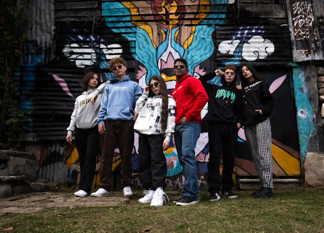 A group of teenagers confidently posing in front of a vibrant, artistic mural, each dressed in casual streetwear. The image captures the fun and creativity that DIY quinceañera choreography can bring when guided by professional resources, emphasizing individuality and teamwork among the court members.