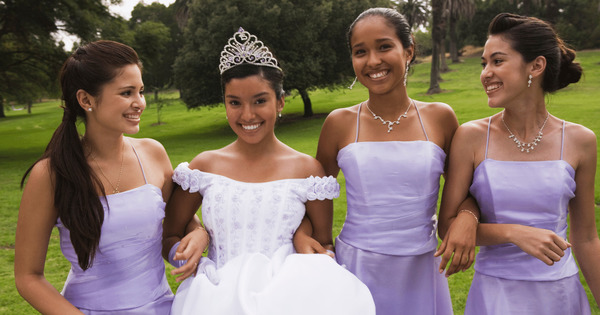  A quinceañera in a beautiful white gown and tiara smiles while standing arm-in-arm with her damas, who are dressed in matching lavender gowns. The image highlights the significance of choosing close friends or family members for your quinceañera court to create a supportive and joyful group.