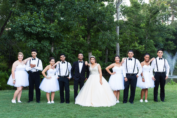 A quinceañera in a stunning white gown stands with her court on a lush green lawn. The chambelanes are dressed in black suits with white shirts, black suspenders, and bow ties, while the damas wear short white dresses. The group poses confidently, showcasing the importance of selecting a well-coordinated quinceañera court.