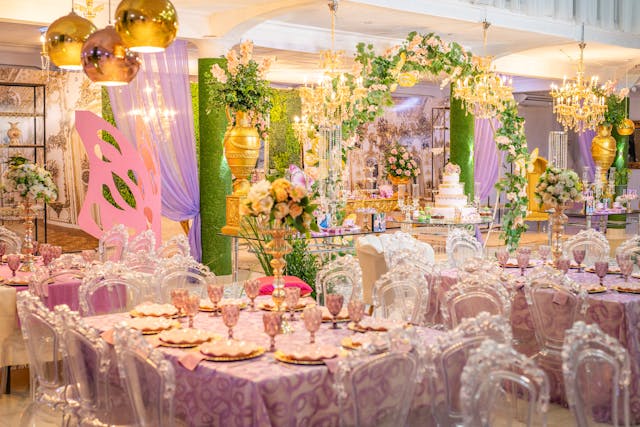 A grand reception hall set with beautifully decorated tables featuring purple tablecloths, crystal-clear chairs, and elaborate floral centerpieces. Chandeliers and draped greenery create a magical setting for a quinceañera reception, ensuring a flawless event experience.