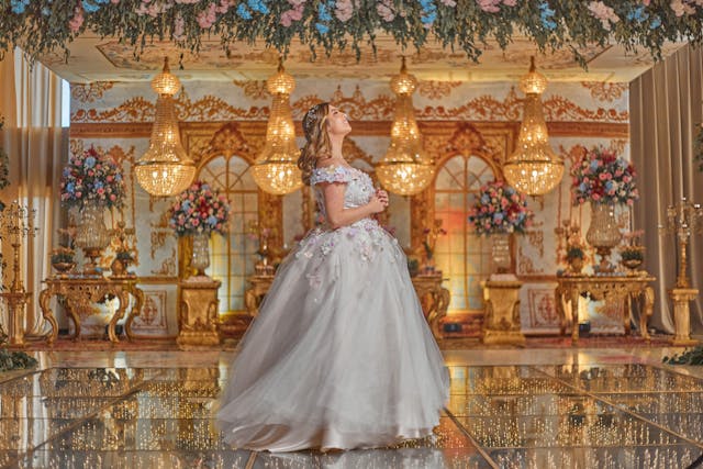 A quinceañera in a stunning gown stands gracefully on a reflective dance floor under elegant chandeliers, surrounded by luxurious décor. The image captures a key moment in the reception timeline, emphasizing the beauty and sophistication of the event.