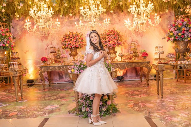 A smiling quinceañera poses in a short, detailed white dress in front of an ornate dessert table adorned with chandeliers, flowers, and colorful decorations. The image highlights the celebratory atmosphere and importance of key moments during the quinceañera reception.