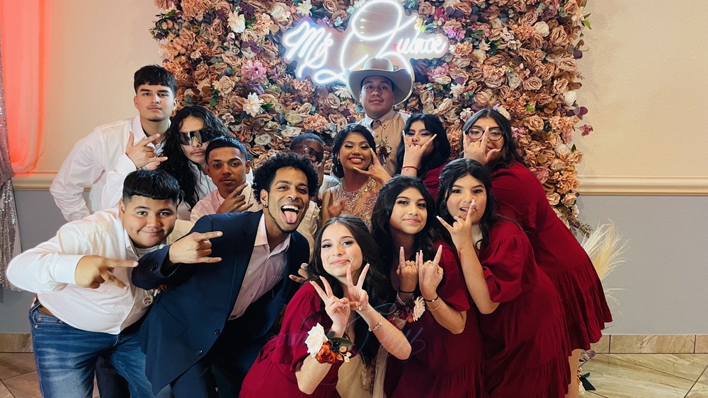 A quinceañera and her court of honor pose playfully in front of a flower wall backdrop with a neon sign. The group, dressed in coordinated outfits, smiles and makes fun hand gestures, capturing the joy and camaraderie that comes with preparing for a quinceañera celebration.