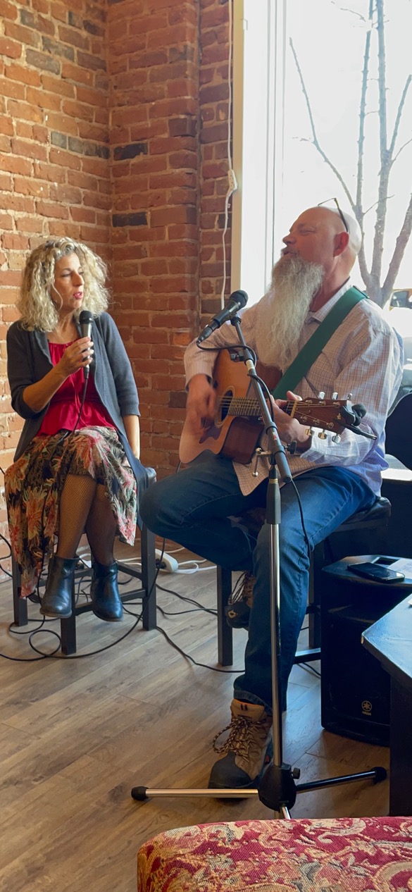 John and Monique Dingeldein performing live at the Old Trolley Barn in Spokane. John, with a long white beard, is playing an acoustic guitar while Monique sings into a microphone. They are seated against a rustic brick wall near a window, creating a cozy and intimate atmosphere for the audience. John and Monique Dingeldein performing live at the Old Trolley Barn in Spokane. John, with a long white beard, is playing an acoustic guitar while Monique sings into a microphone. They are seated against a rustic brick wall near a window, creating a cozy and intimate atmosphere for the audience.