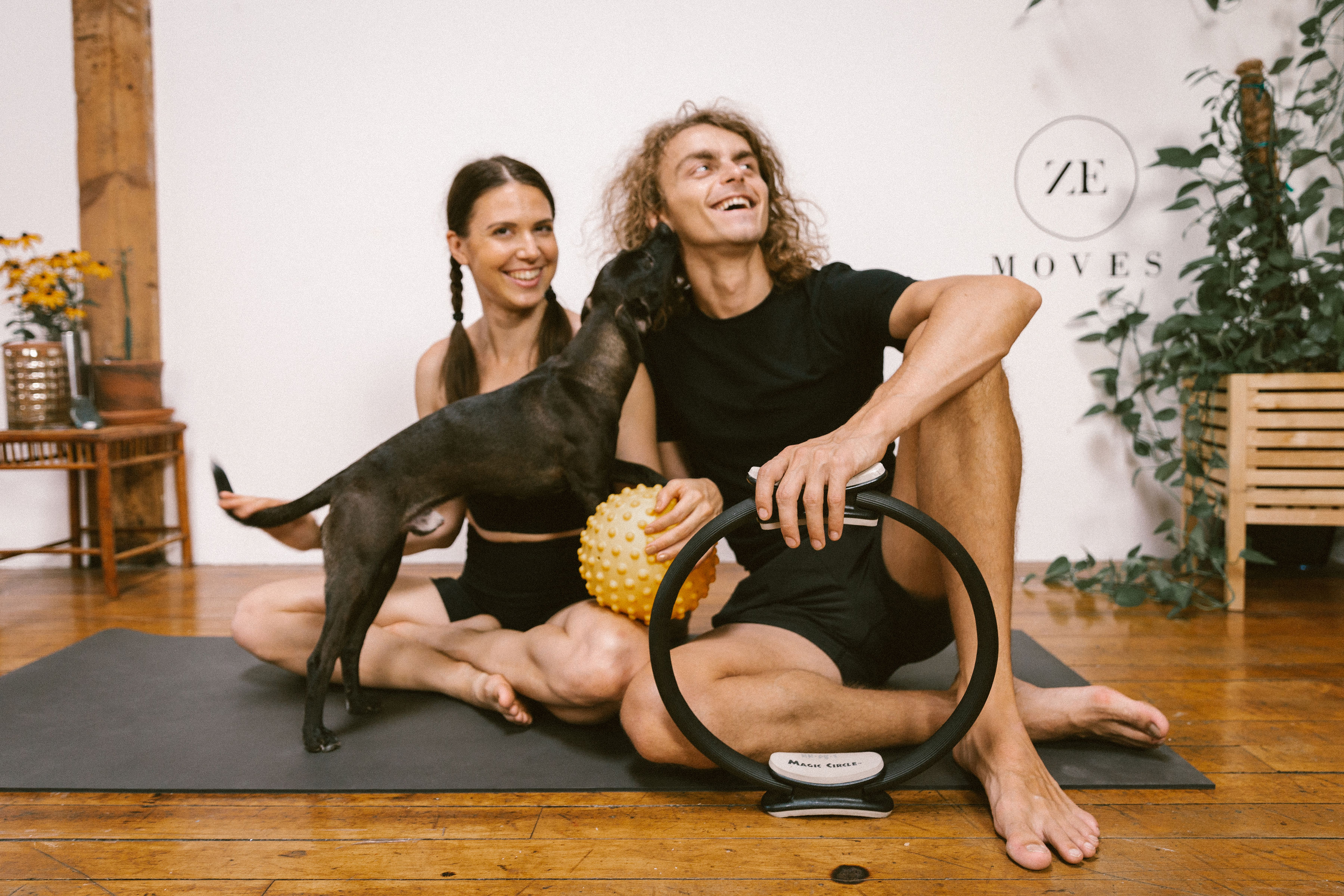 A couple seated on the floor with a pilates circle and ball with a small black dog.