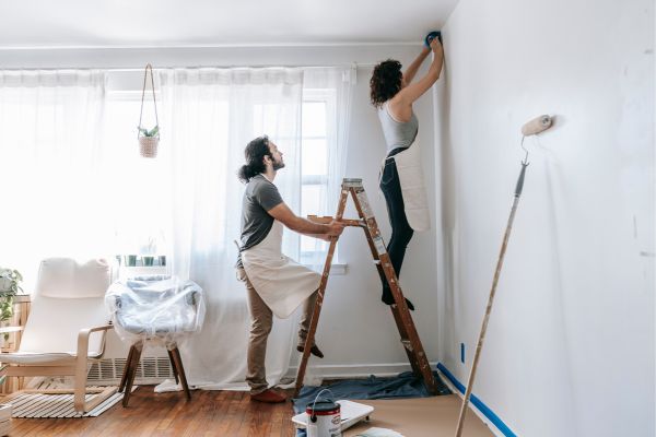 Couple prepping wall for interior painting in living room with ladder and paint tools.jpg Couple prepping wall for interior painting in living room with ladder and paint tools.jpg