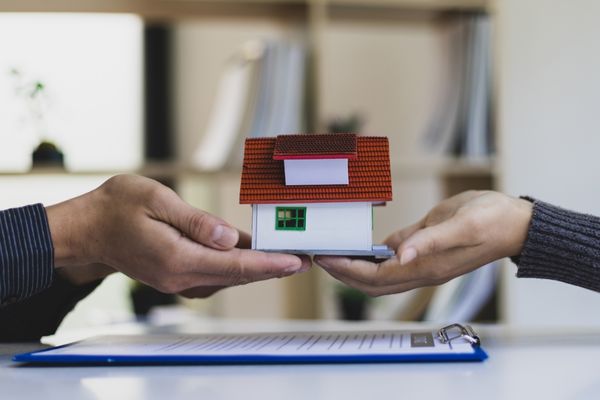 Two hands holding a miniature house model over a contract clipboard Two hands holding a miniature house model over a contract clipboard