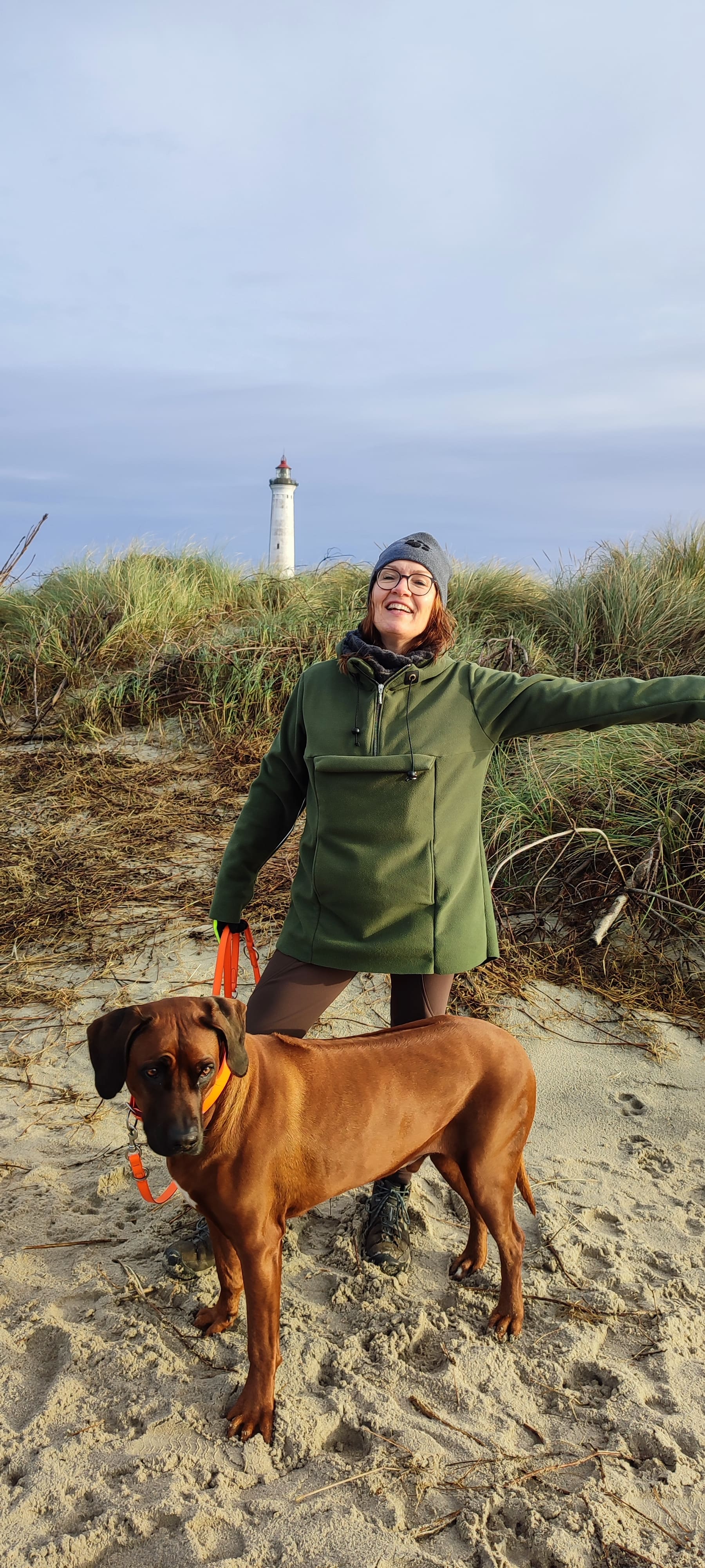 Katja steht lachend mit ihrem Hund am Strand vor dem Leuchtturm Lyngvig Fyr. Sie trägt einen selbstgenähten grünen Anorak – Symbol für Kreativität, Erdung und Selbstwirksamkeit.