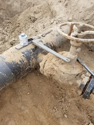 A partially unearthed metal pipe with a valve is shown embedded in dry, sandy soil. The pipe appears corroded and is exposed for manual access, possibly for inspection or repair of a joint or damaged section.