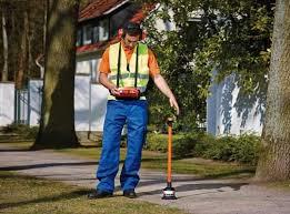 A worker in a safety vest and hard hat uses an acoustic resonating locator to scan the ground along a sidewalk near trees and residential homes. The locator device is held vertically and appears to be detecting underground non-metallic water lines such as PVC by sound.