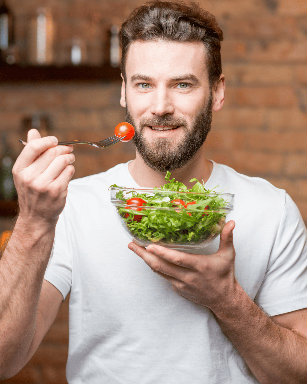A smiling man with a beard wearing a white t-shirt holds a bowl of fresh salad with cherry tomatoes and leafy greens while lifting a forkful to eat. Represents healthy eating and an anti-inflammatory diet. A smiling man with a beard wearing a white t-shirt holds a bowl of fresh salad with cherry tomatoes and leafy greens while lifting a forkful to eat. Represents healthy eating and an anti-inflammatory diet.