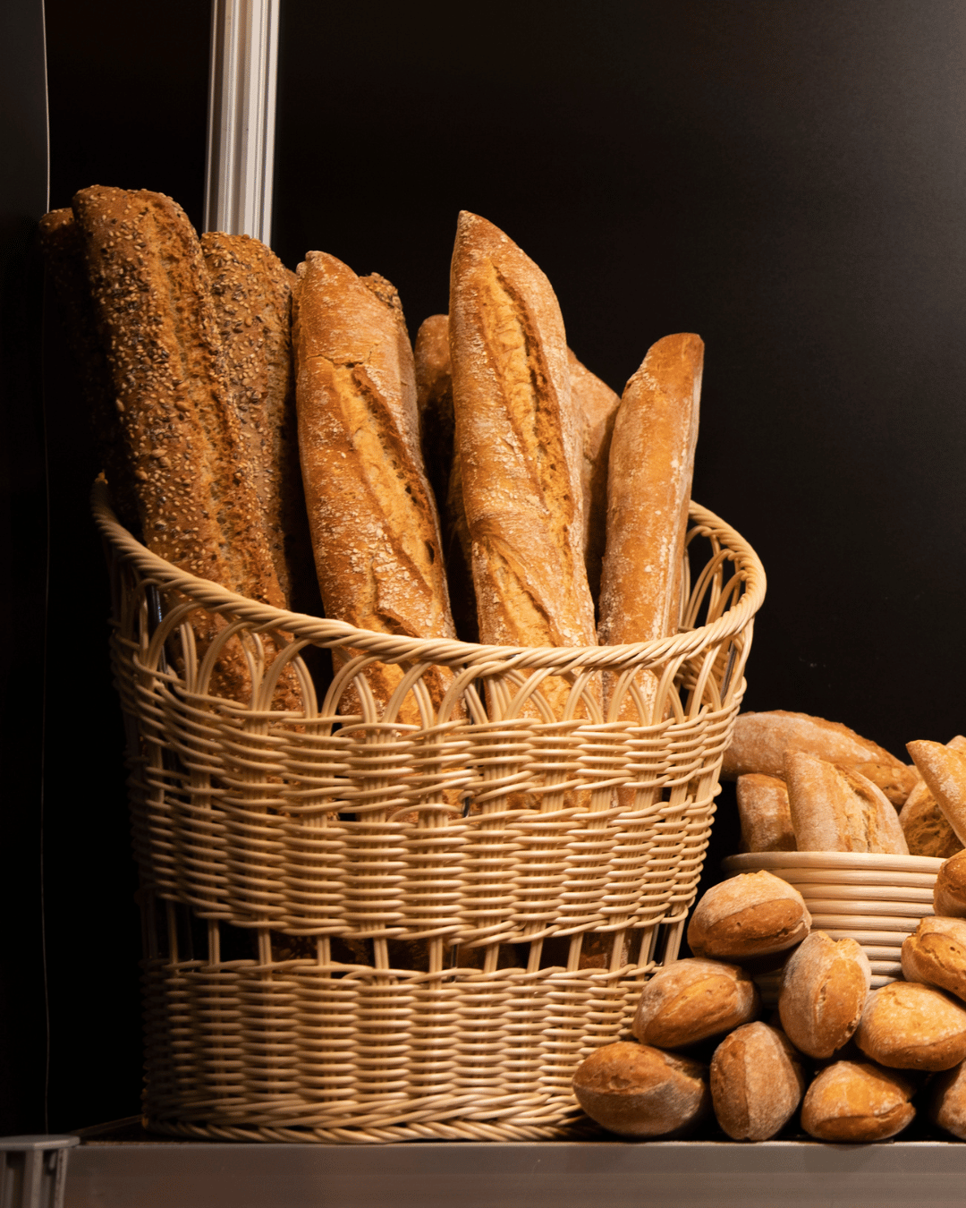 A basket filled with freshly baked baguettes and whole grain bread, surrounded by small artisan rolls, against a dark background. Represents gluten, wheat, and traditional bakery products. A basket filled with freshly baked baguettes and whole grain bread, surrounded by small artisan rolls, against a dark background. Represents gluten, wheat, and traditional bakery products.