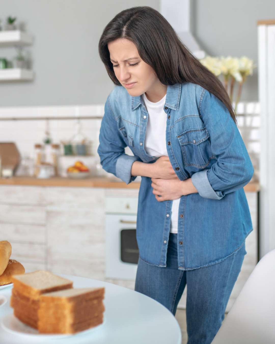 Woman clutching her stomach in discomfort while standing near a table with bread, representing digestive distress often associated with non-celiac gluten sensitivity (NCGS). Woman clutching her stomach in discomfort while standing near a table with bread, representing digestive distress often associated with non-celiac gluten sensitivity (NCGS).