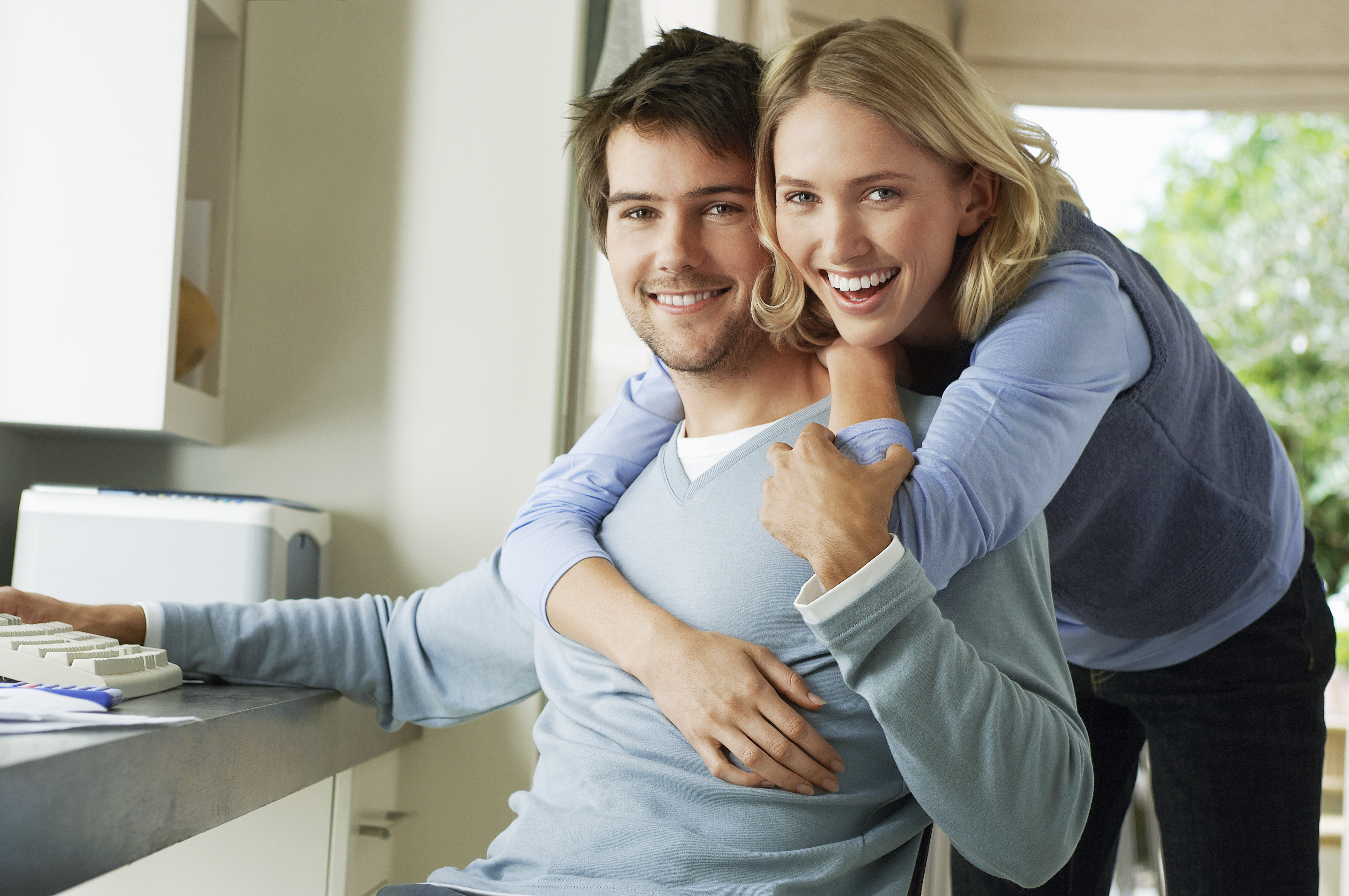 man sitting at desk and wife hugging him from behind. They are both smiling at the camera