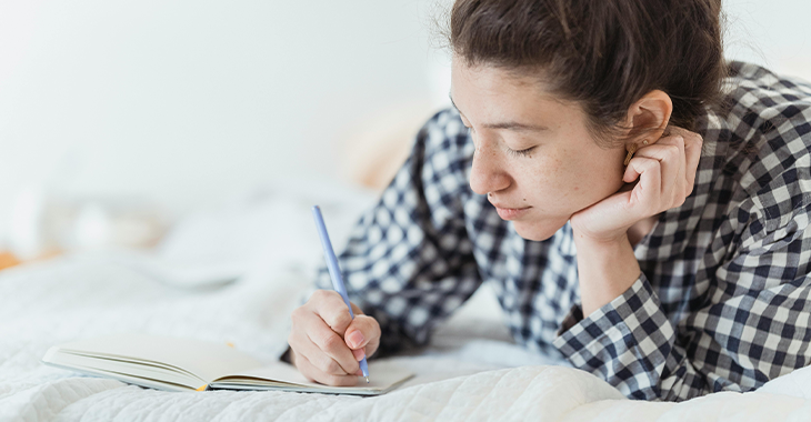 Woman lying on a bed, writing in her journal, reflecting on her emotions and practicing stress management through journaling. Woman lying on a bed, writing in her journal, reflecting on her emotions and practicing stress management through journaling.