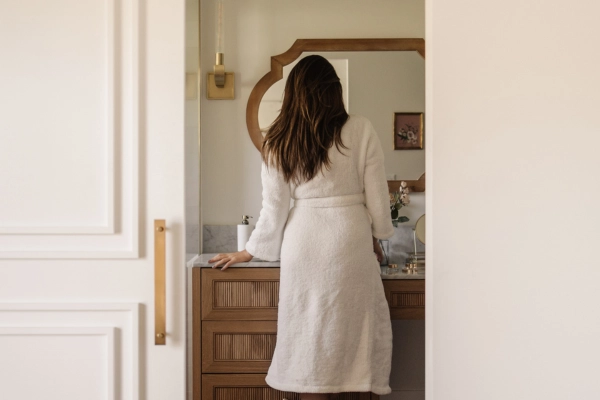 Woman in white bathrobe standing at wooden bathroom vanity looking in round mirror, creating a serene morning wellness routine