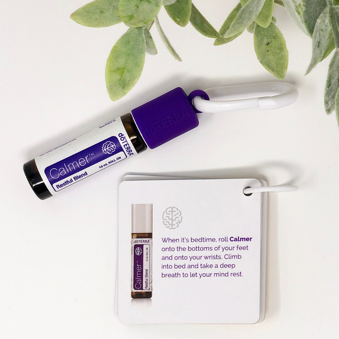 A photo of a roller bottle filled with doTERRA's Calmer blend of essential oils for kids lying on a white surface next to a flashcard that describes the benefits of using Calmer for bedtime without tears or tantrums