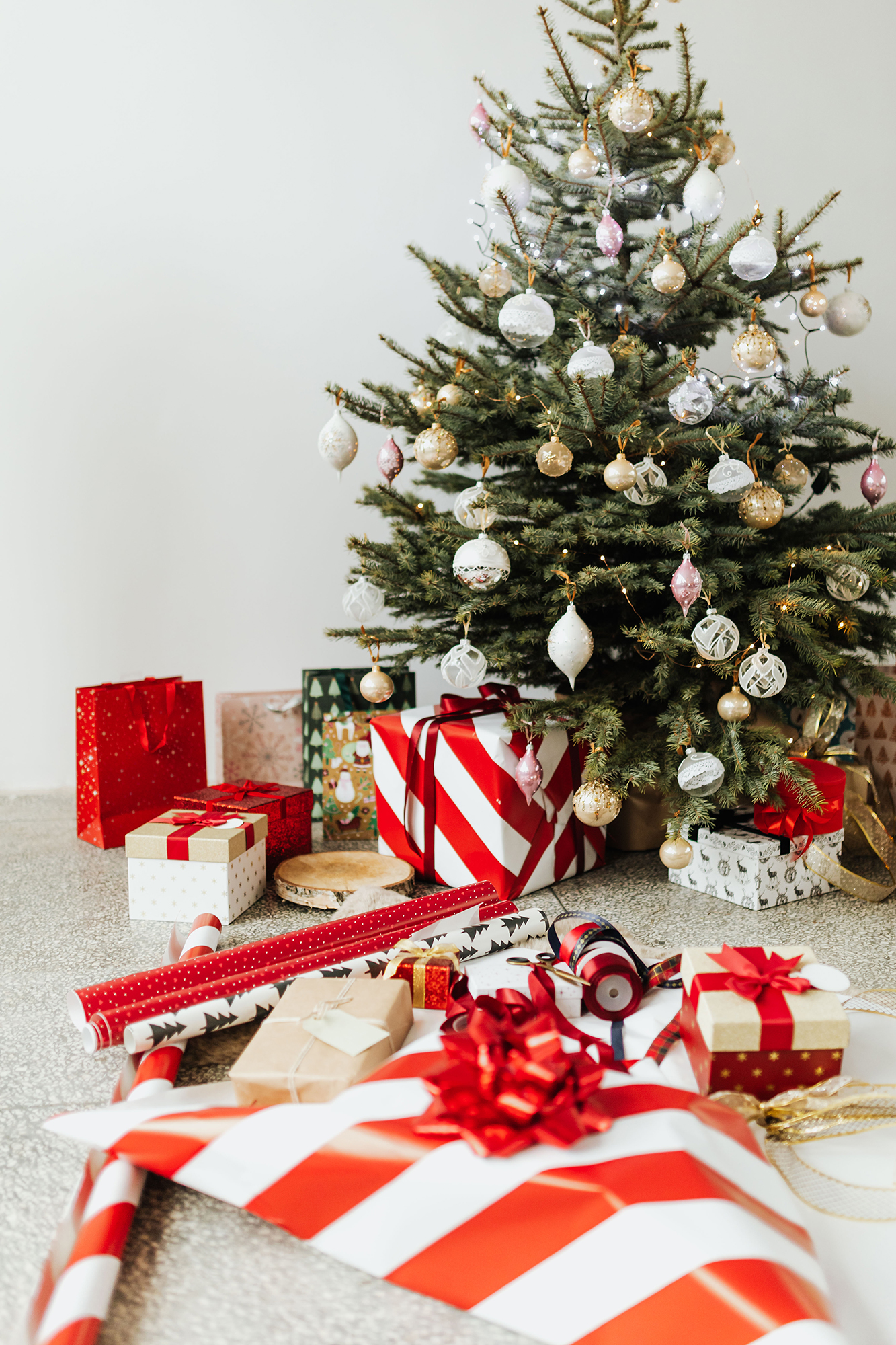 Photo of a Christmas tree surrounded by gift-wrapped presents and wrapping paper, bows, and ribbons. The tree is decorated with ornaments and ready for the holidays