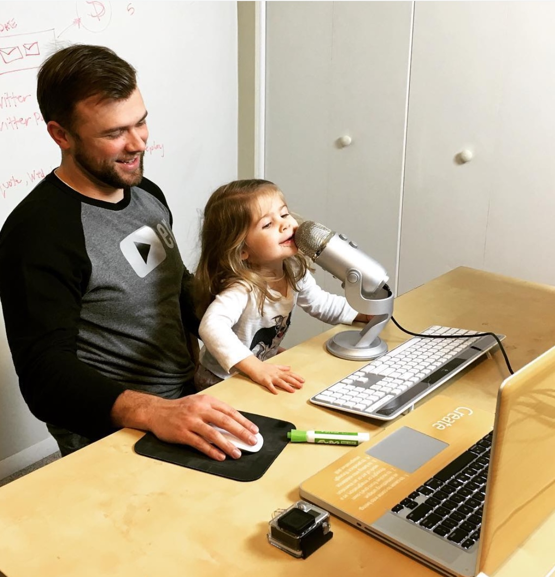 Jake Larsen sits at his desk, smiling as his young daughter enthusiastically speaks into a microphone. A laptop, keyboard, and other work essentials are on the desk, while a whiteboard filled with notes is visible in the background. This moment captures the blend of entrepreneurship and family life, highlighting the early days of building Video Power Marketing from home.