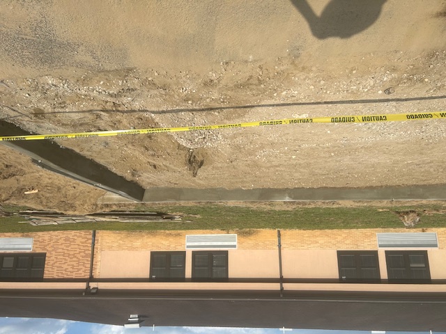 Partially constructed or unrenovated railroad industrial site with a long, narrow concrete trench running through the center. The trench is bordered by raised concrete edges, and the surrounding area appears to be a warehouse or a similar large indoor space. Scaffolding, construction equipment, and safety barriers are visible in the background, indicating ongoing construction or maintenance work.