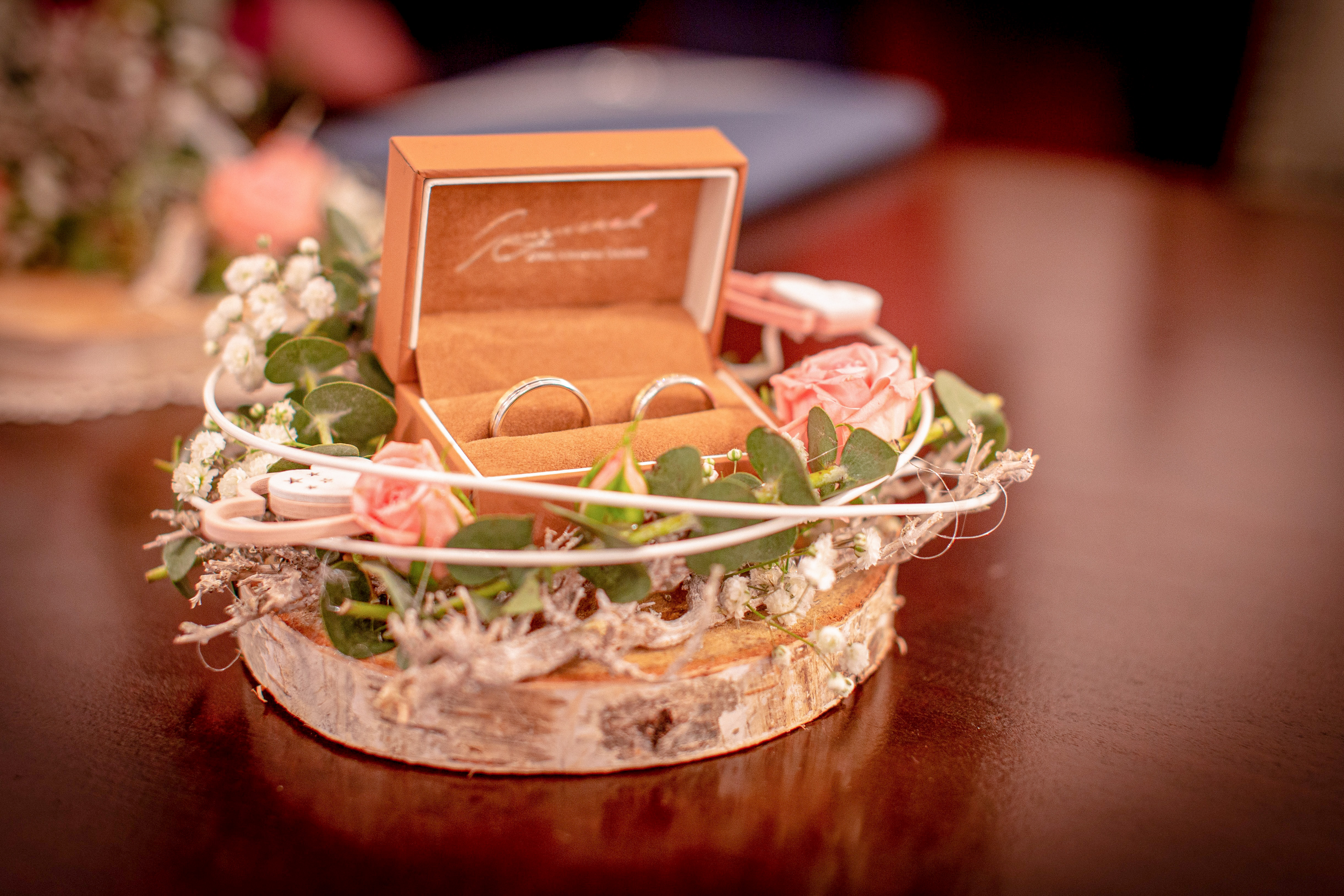 two rings in a light brown ring box on a slice of wood with flowers around it