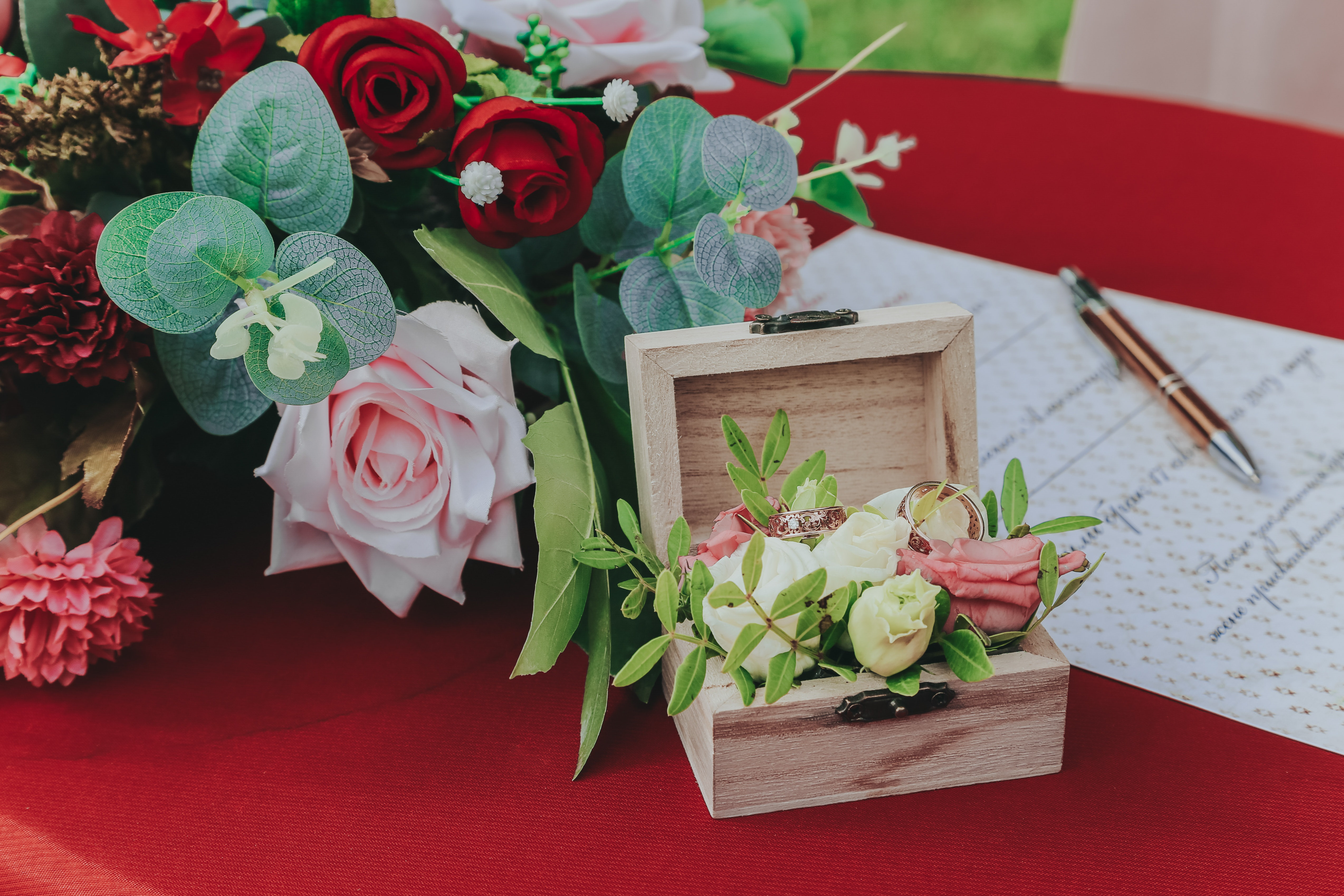 red table cloth holds wooden wood box and bridal bouquet while the wedding bands sit on top of flowers in the ring box