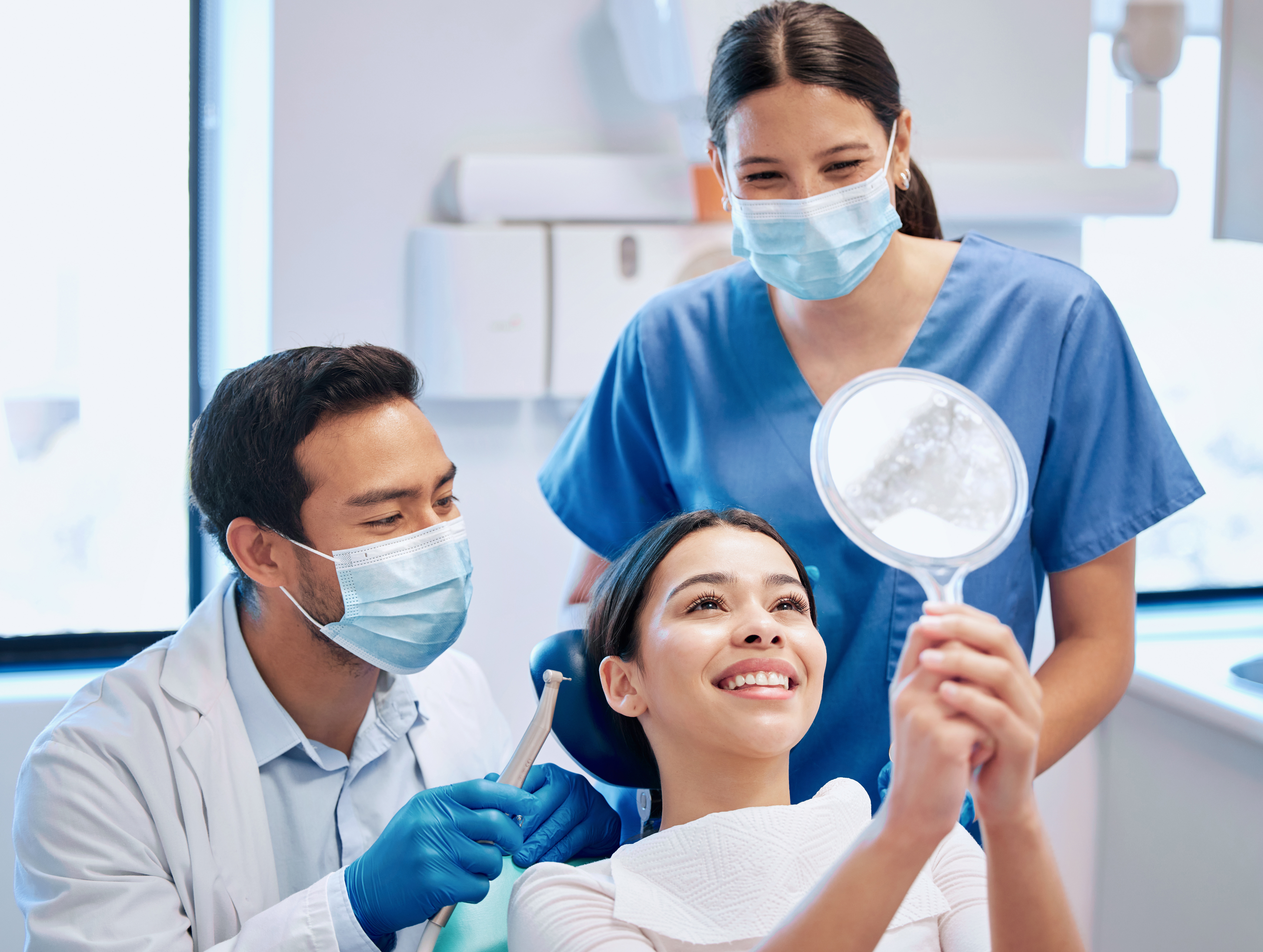 Dentist marketing photo of a happy patient smiling with a mirror after treatment, assisted by a dentist and dental assistant in a modern clinic.