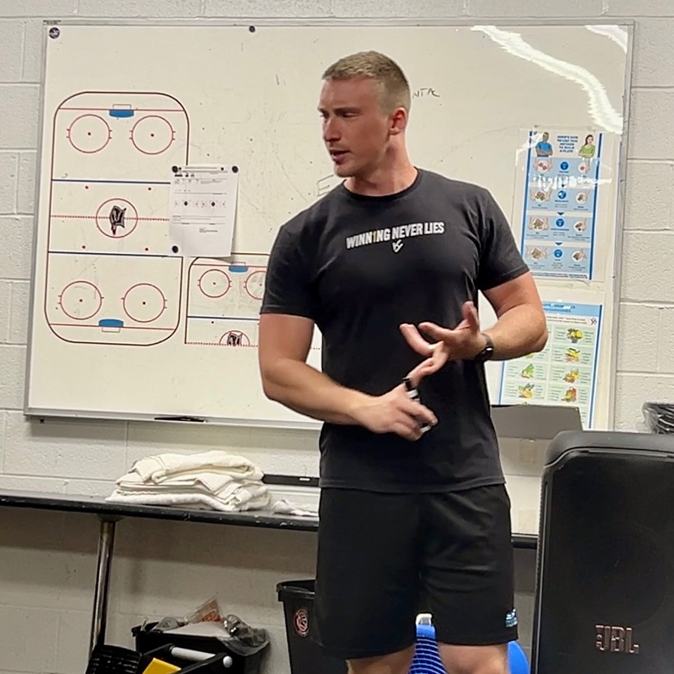 Hockey coach in black “Winning Never Lies” shirt giving a motivational team talk in locker room with whiteboard rink diagrams.