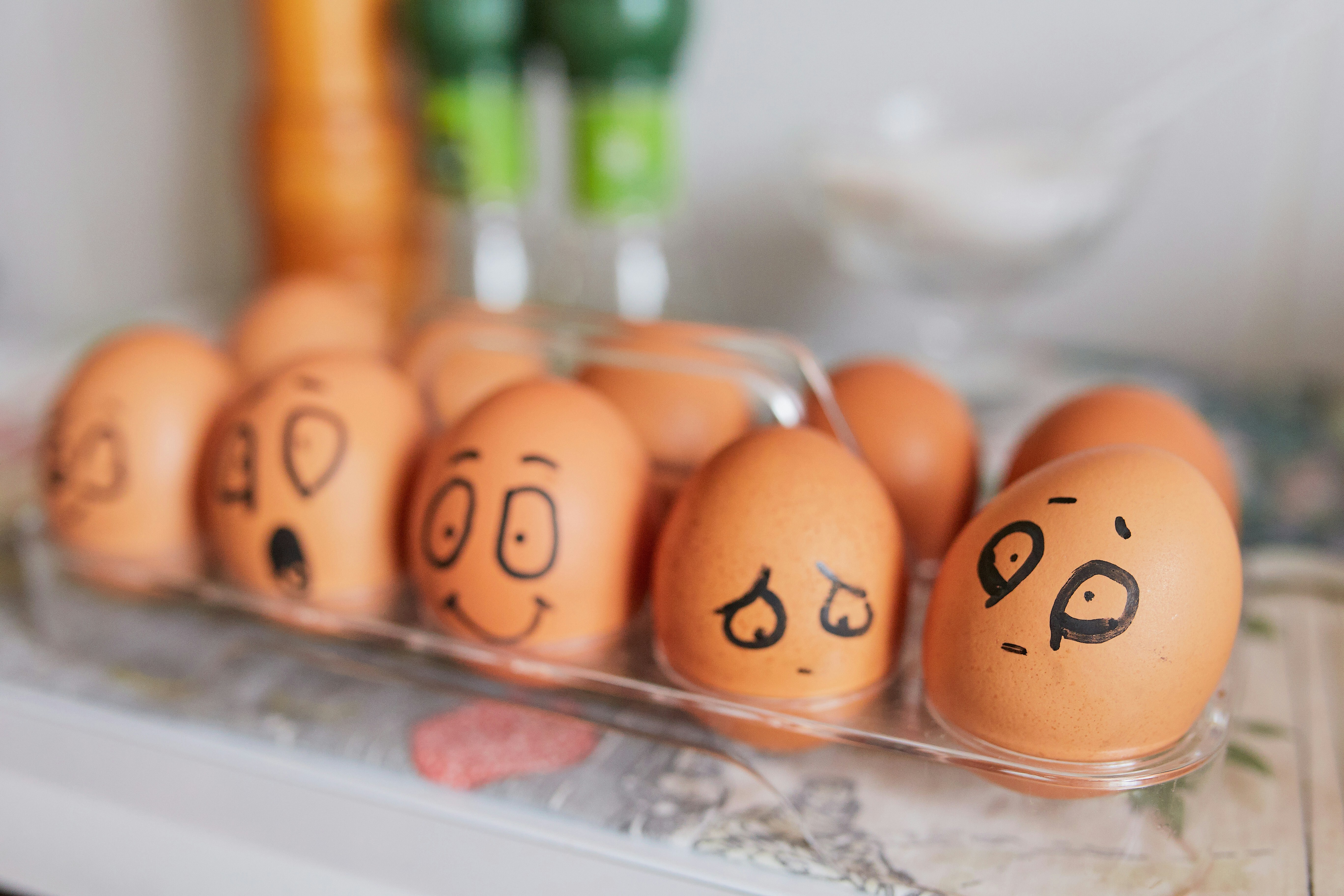 A group of brown eggs in a transparent carton, each with different hand-drawn facial expressions, including happy, sad, surprised, and worried, creating a humorous and expressive scene in a kitchen setting A group of brown eggs in a transparent carton, each with different hand-drawn facial expressions, including happy, sad, surprised, and worried, creating a humorous and expressive scene in a kitchen setting